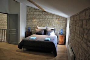 A bedroom with a stone wall, a bed with pillows, a nightstand with a lamp, and a door.
