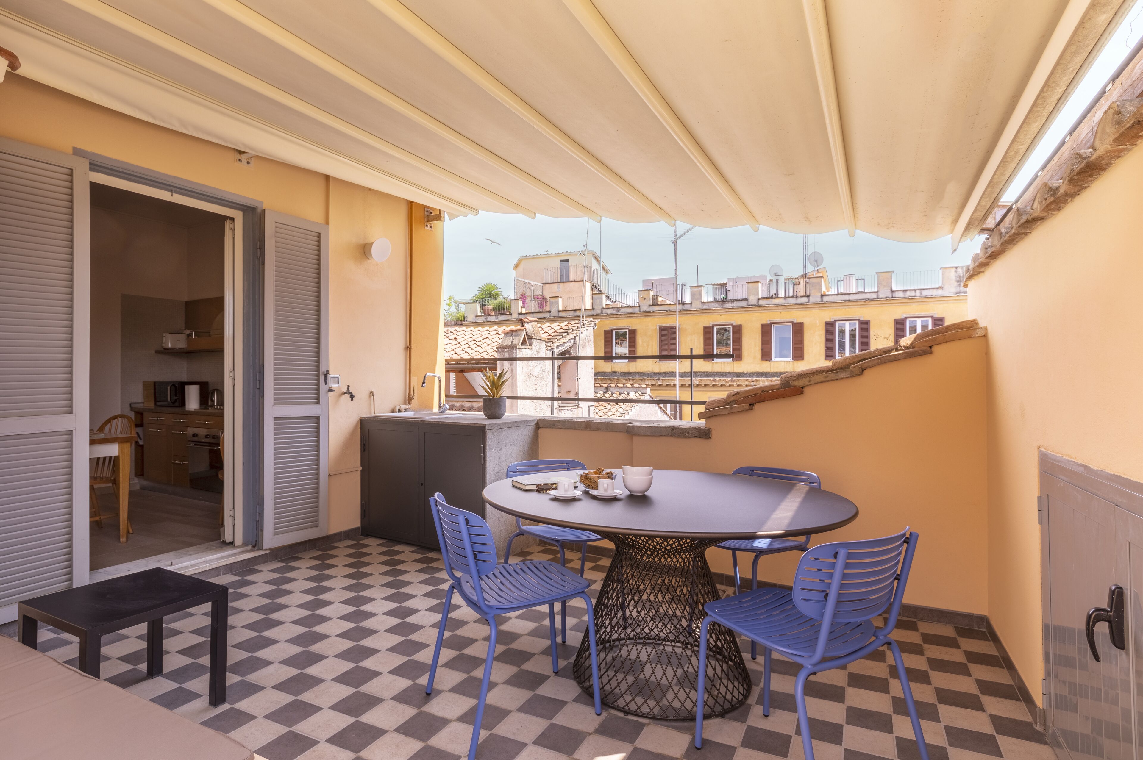 A balcony with a checkered floor, a round table with chairs, and a view of buildings in the background.