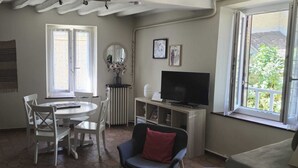 Ein Zimmer mit einem runden Esstisch, weißen Stühlen, einem Fernseher auf einem Holzständer und einem Fenster mit Blick auf Grünflächen.