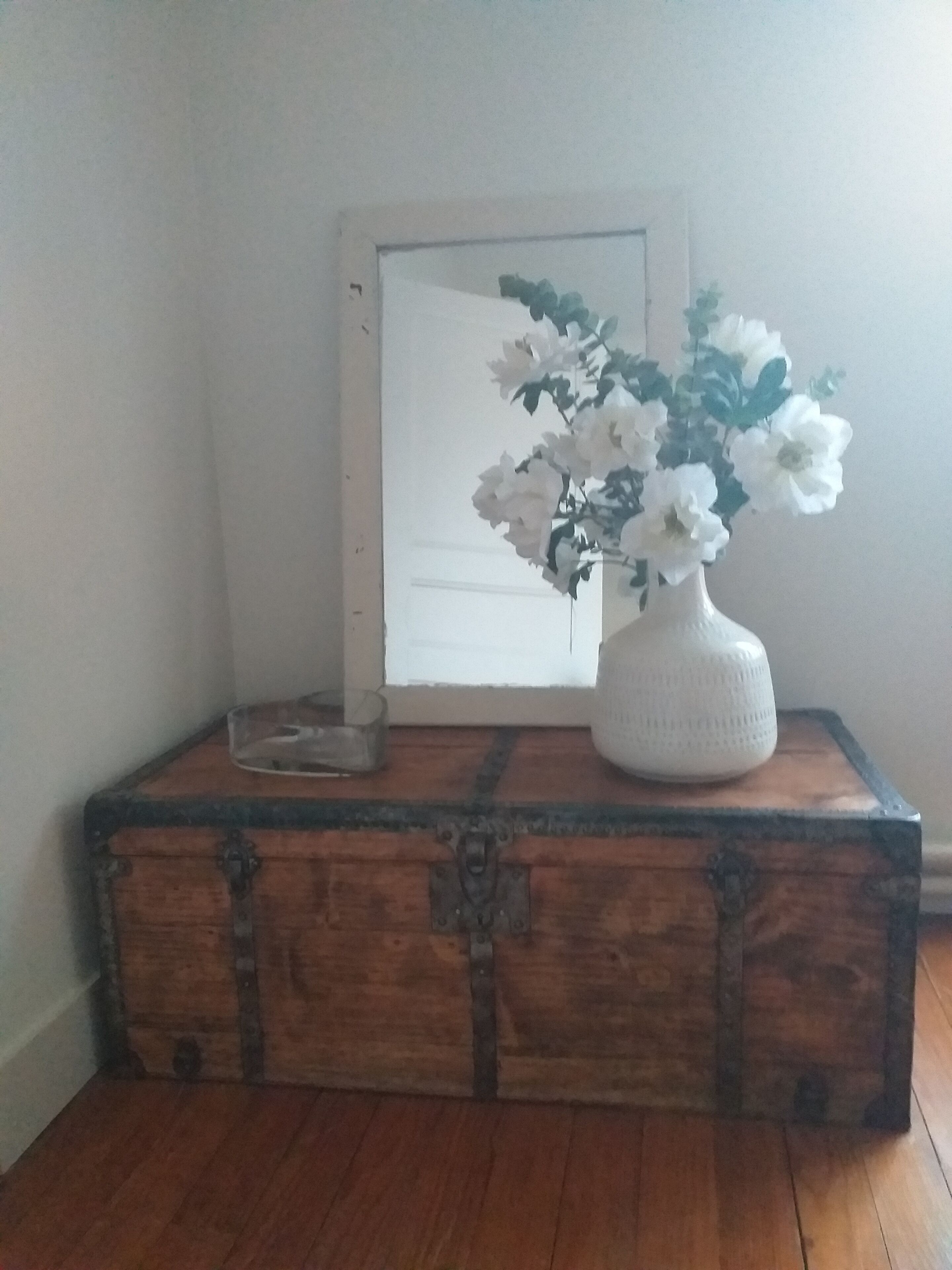 Een houten kist met een vaas witte bloemen erop, geplaatst op een houten vloer naast een witte deur.