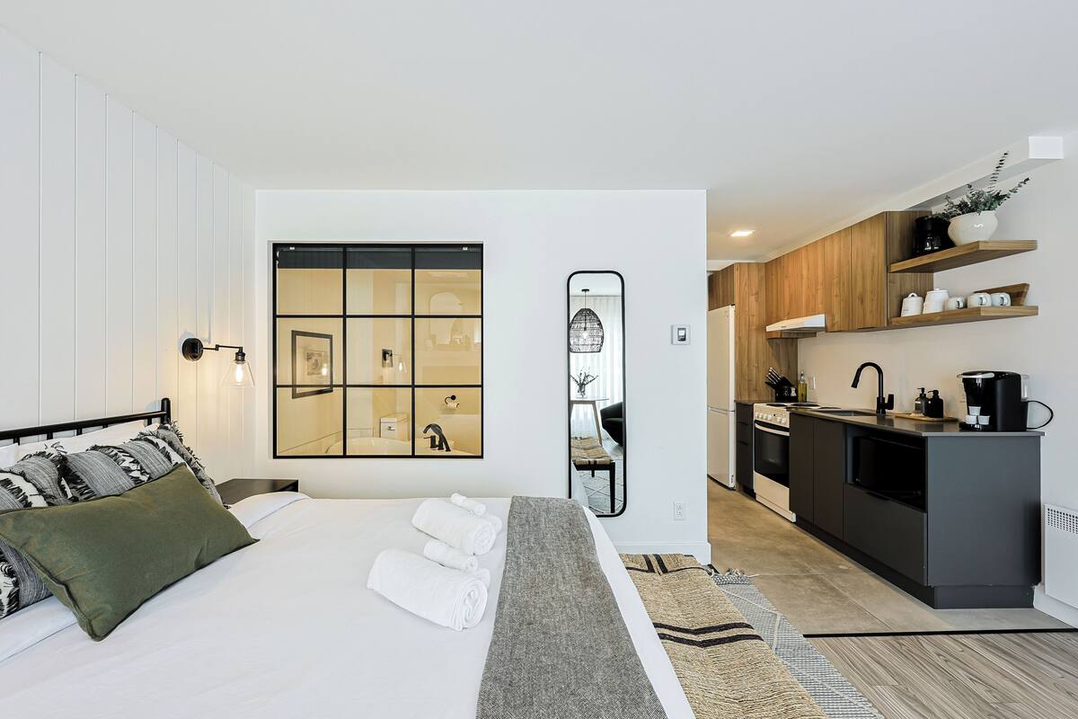 A modern hotel room with a bed, a large mirror, a small kitchenette, and a view of the city.