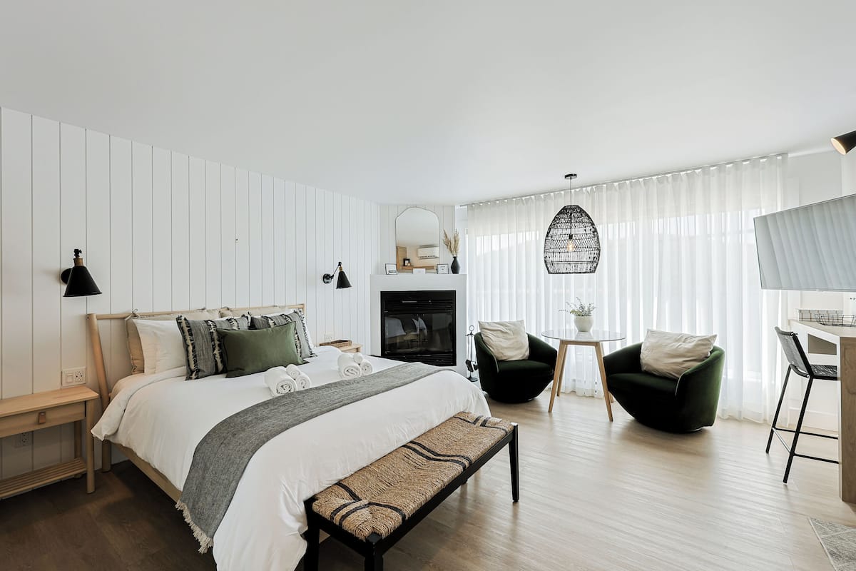 A modern bedroom with a bed, bedside tables, a bench, a chair, a desk, a mirror, a hanging light, and a fireplace.