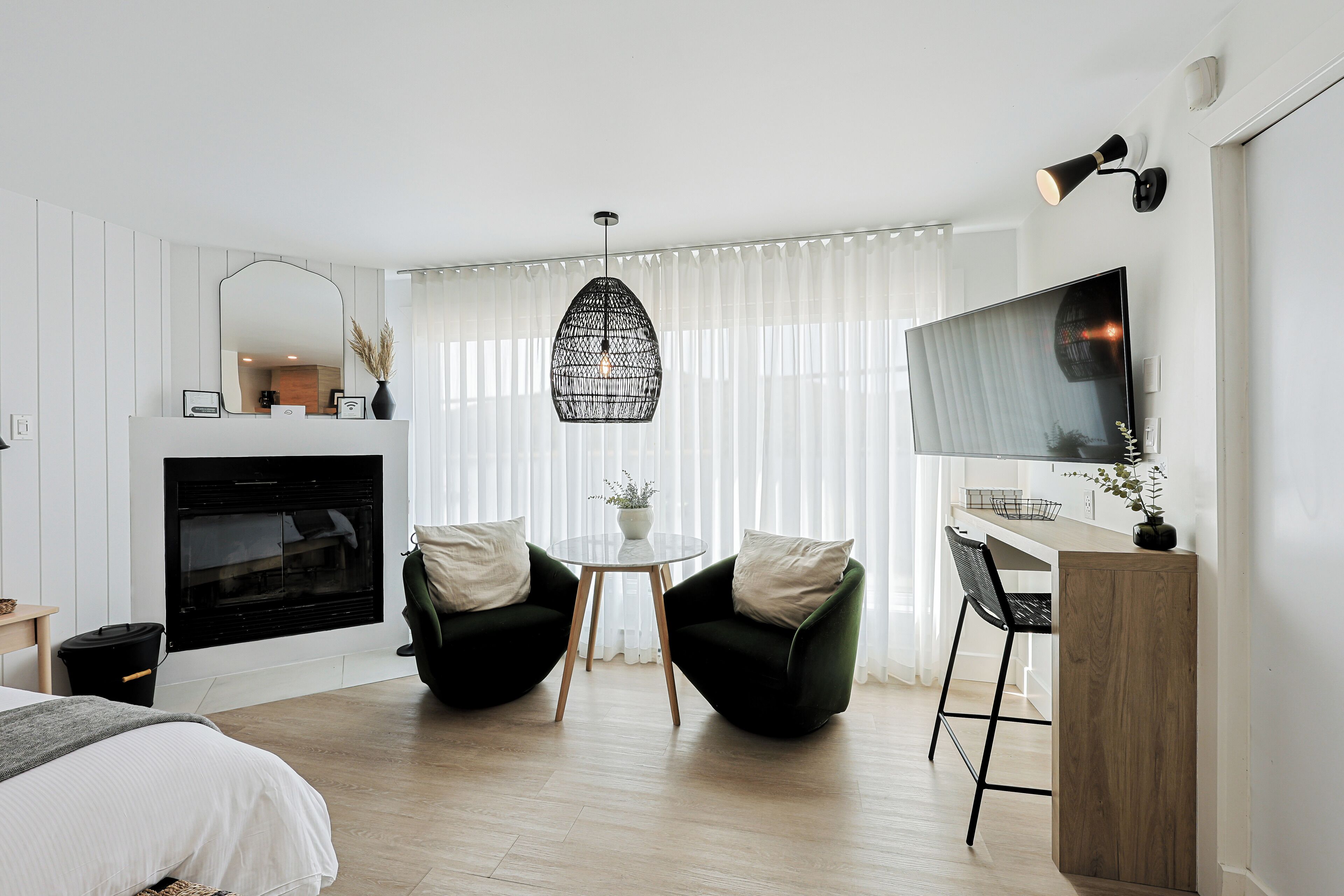 A modern hotel room with a bed, a fireplace, two chairs, a small table, and a TV mounted on the wall.