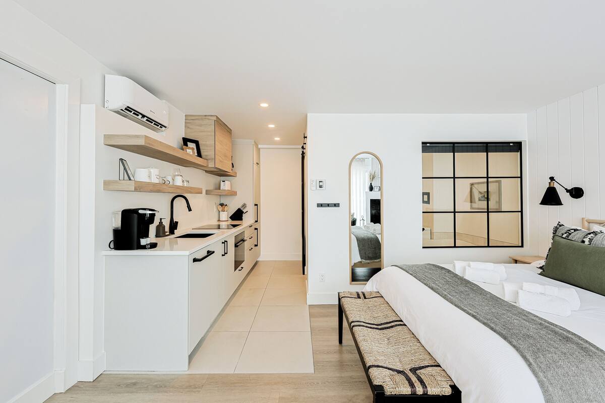 A modern hotel room with a kitchenette, a large bed with a bench, a mirror, and a window with a view.