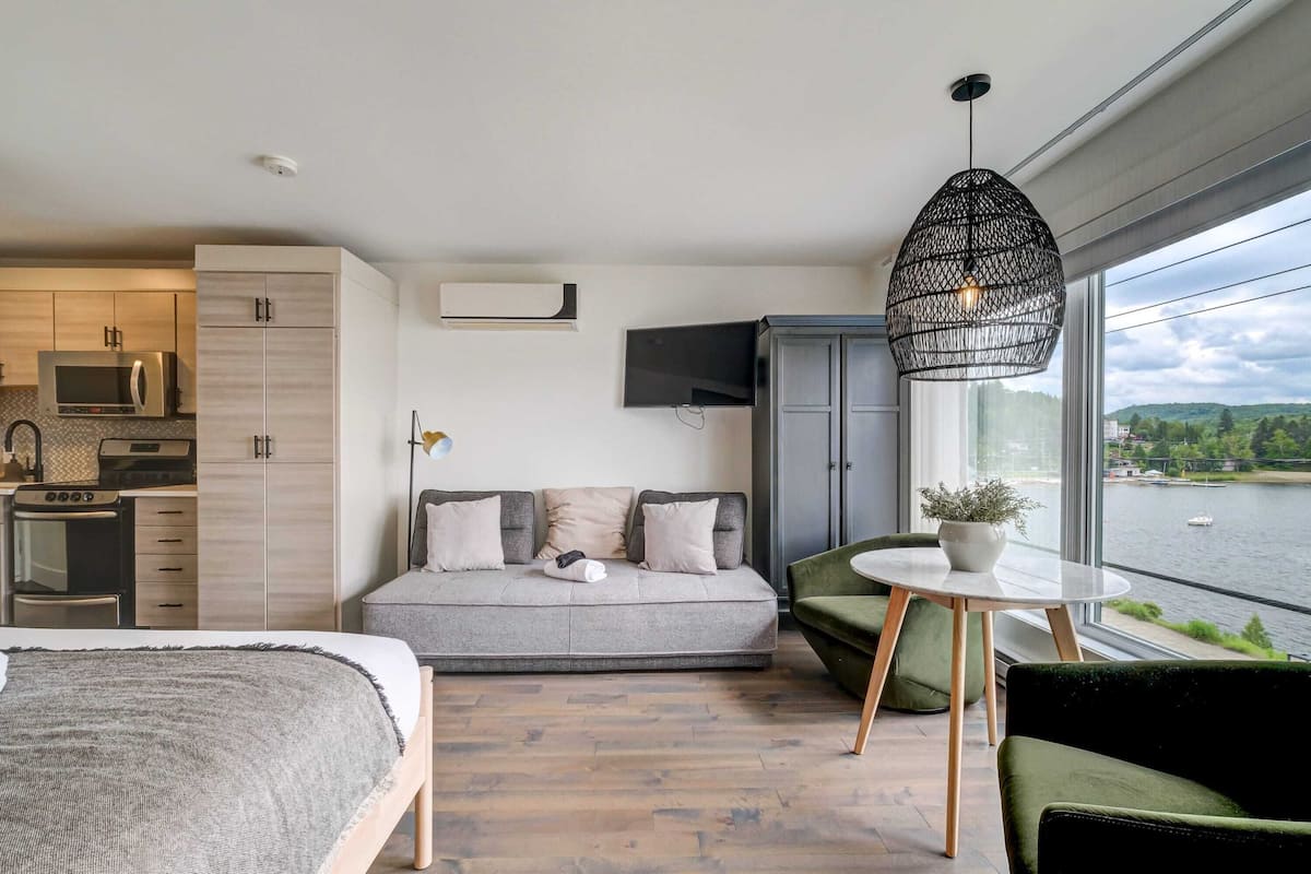 A modern hotel room with a large bed, a sofa, a dining table, and a view of a body of water.
