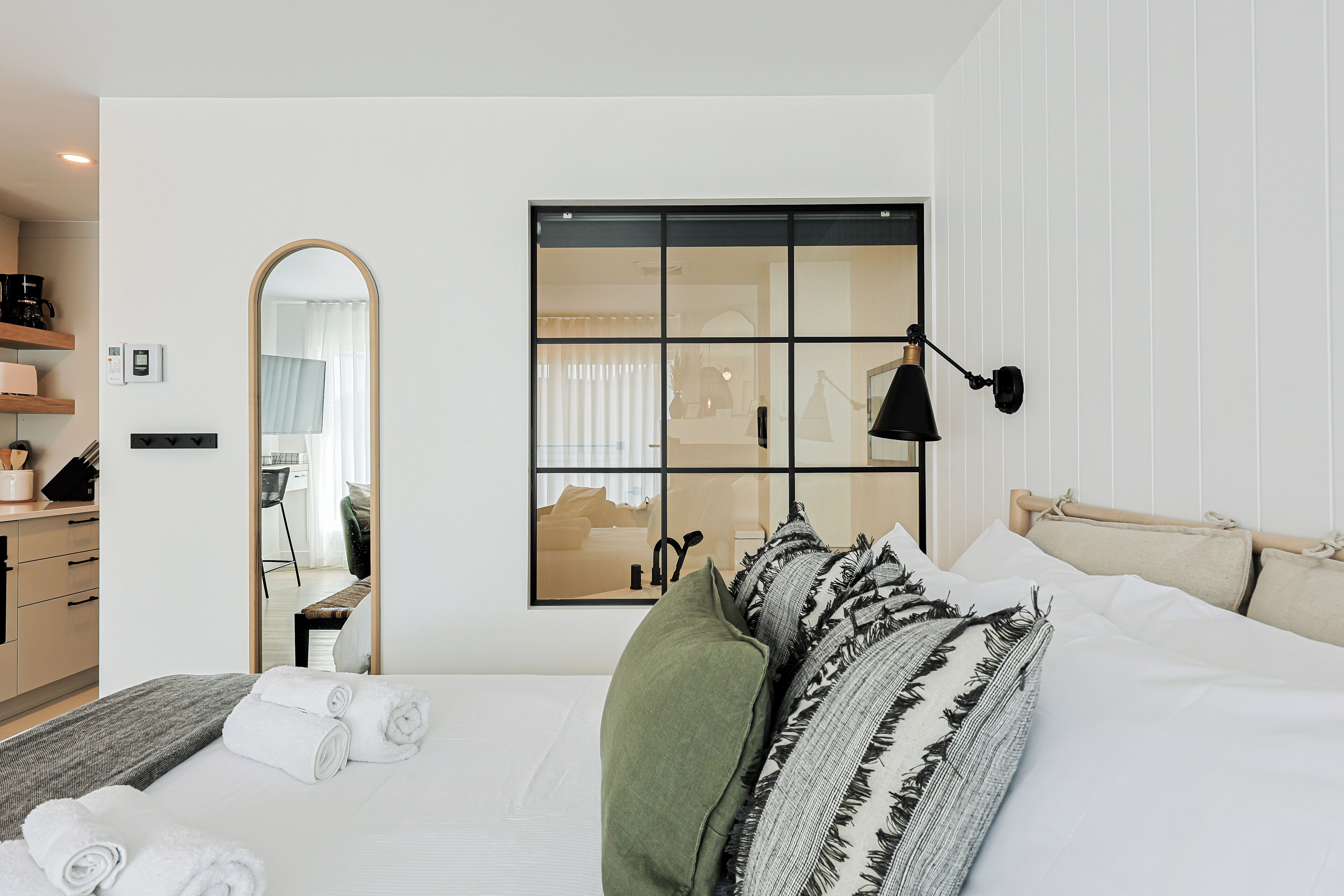 A hotel room with a bed, a large mirror, a window with blinds, a bedside lamp, and a small table with a coffee maker.