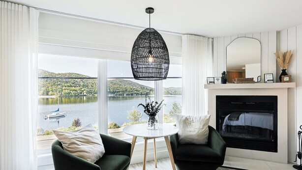 Een moderne woonkamer met uitzicht op een meer en heuvels, met twee groene fauteuils, een ronde tafel en een open haard.