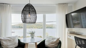 Een moderne kamer met een groot raam, een ronde tafel, twee groene fauteuils, een hanglamp en een flatscreen-televisie.