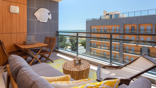 A balcony with a sofa, chairs, and a small table, overlooking a building with multiple windows and a clear view of the sea. - Luxury 2-Bedroom Apartment with RiaView & Rooftop Pool, Village Marina, Olhão (Olhão)