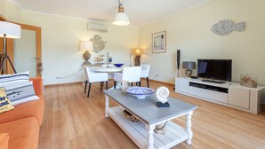 A living room with a sofa, a dining table, chairs, a TV, and a fireplace. - Luxury 2-Bedroom Apartment with RiaView & Rooftop Pool, Village Marina, Olhão (Olhão)
