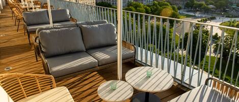 A rooftop terrace with outdoor seating, including sofas and round tables, overlooking a cityscape.