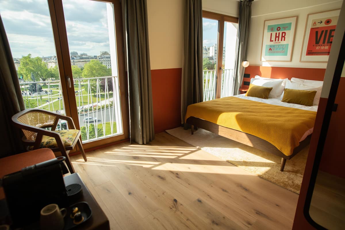 A hotel room with a bed, a chair, a desk, and a view of a cityscape through large windows.