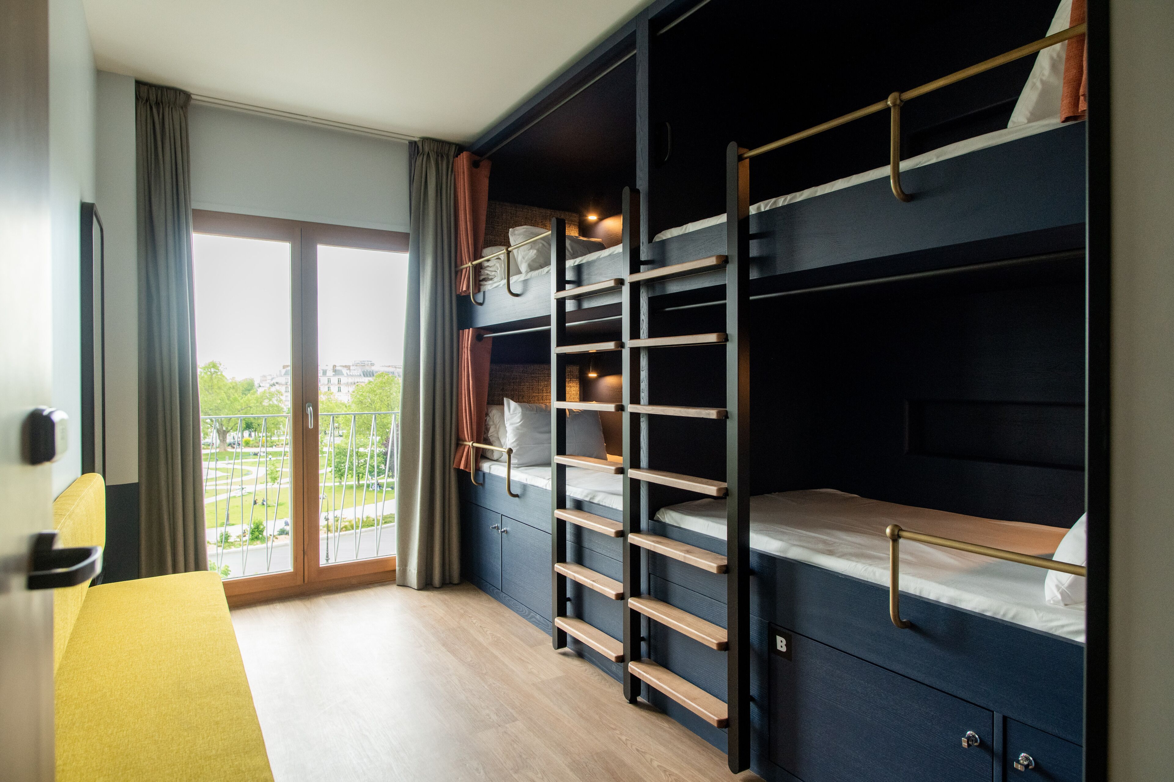 A room with bunk beds, a desk, and a view of a cityscape through a large window.