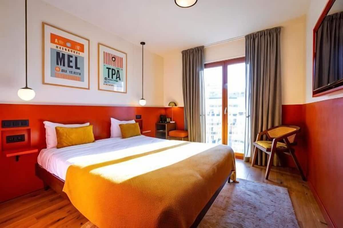 A hotel room with a large bed, a desk, a chair, a window with curtains, and a view of the city.