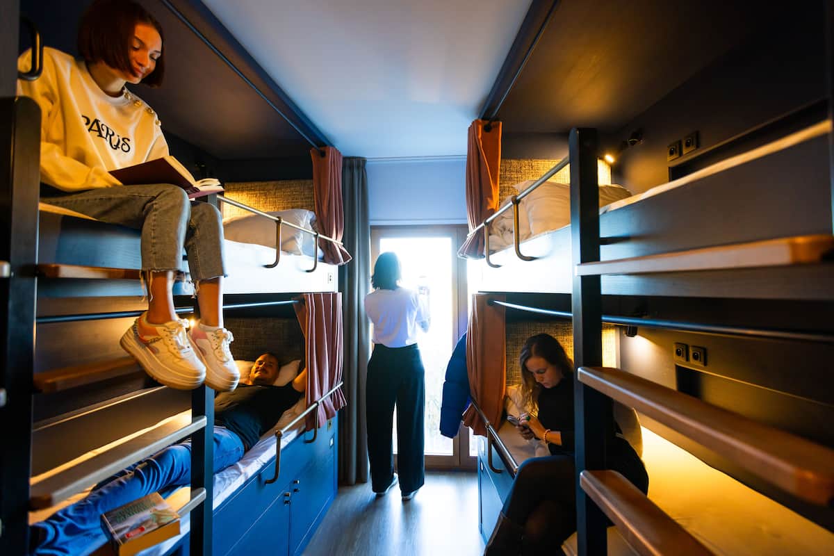 A train compartment with bunk beds, a person reading a book, and another using a mobile phone.