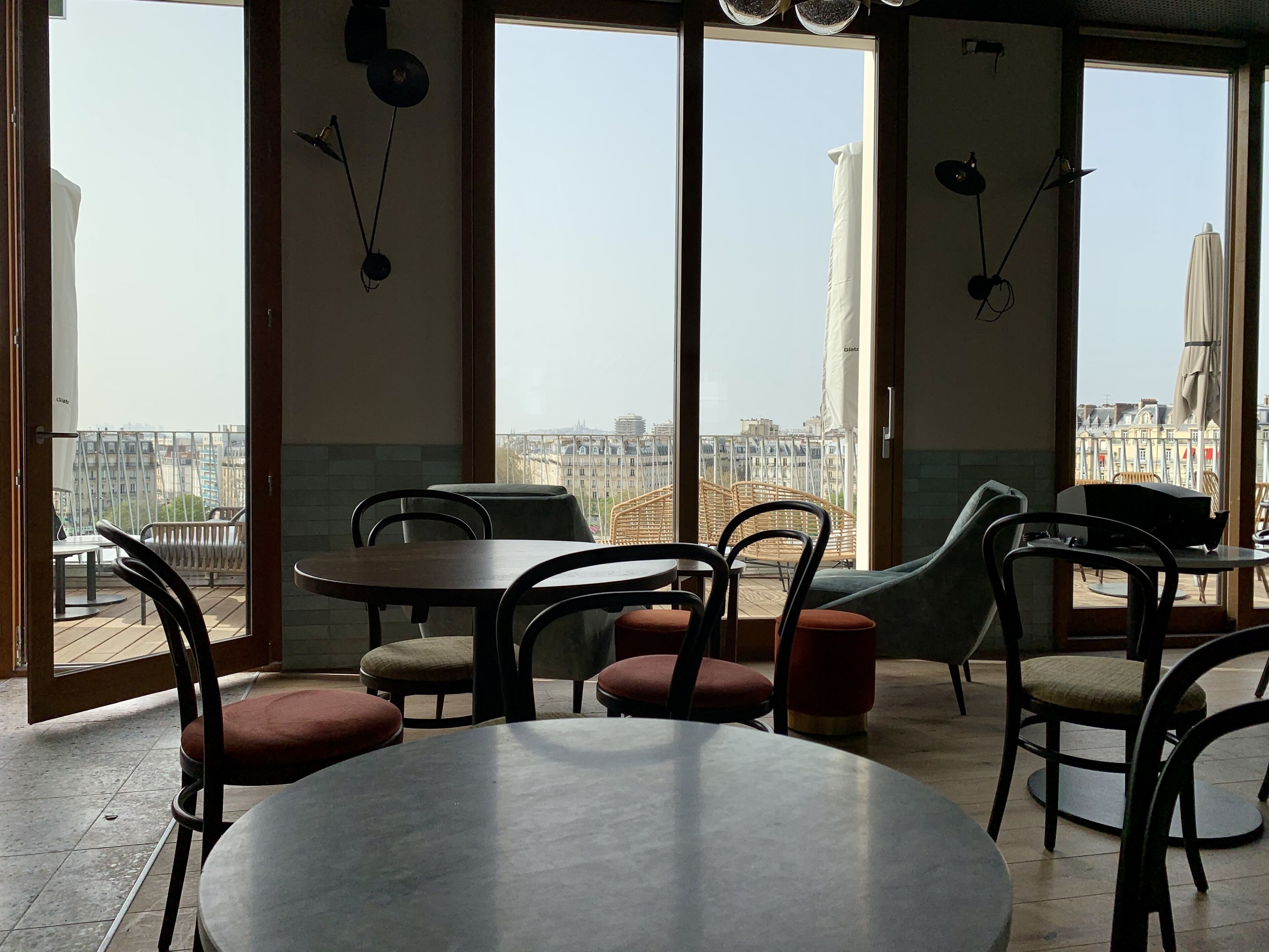 A dining area with round tables and chairs, large windows with views of buildings, and hanging light fixtures.