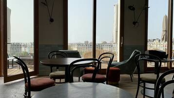 A dining area with round tables and chairs, large windows with views of buildings, and hanging light fixtures.