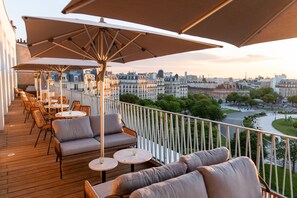Un balcón con mobiliario exterior, incluyendo sofás y mesas, con vistas a un paisaje urbano con edificios históricos.