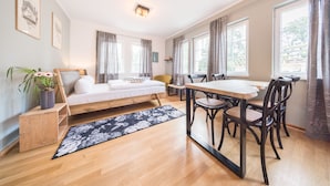 A modern room with a bed, a dining table with chairs, large windows, and a small side table with a plant. - Design Apartments - "Kutscherhaus" (Potsdam)
