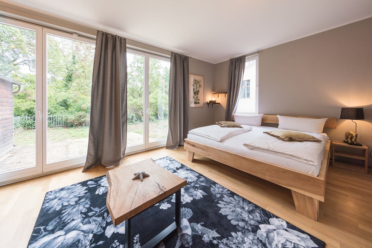 Ein Schlafzimmer mit einem großen Bett, einem Nachttisch aus Holz, einem kleinen Tisch und Blick ins Freie durch große Schiebetüren.