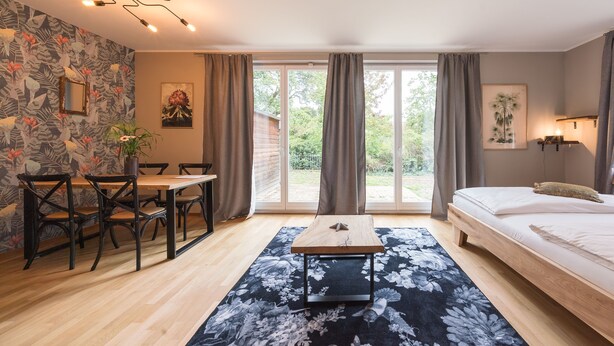 A room with a bed, a dining table with chairs, a small table with a plant, and a large window with curtains. - Design Apartments - "Kutscherhaus" (Potsdam)