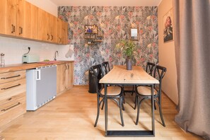 Cuina petita amb armaris de fusta, un frigorífic de dimensions reduïdes, una taula de dinar amb quatre cadires i una paret decorativa amb un motiu floral.