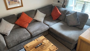 Un salon avec un canapé gris, deux coussins orange et deux coussins gris, une table basse en bois et un tableau encadré accroché au mur.