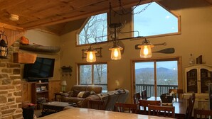 A cozy cabin interior with a stone fireplace, a large TV, a dining area, and a balcony with a mountain view.
