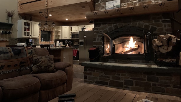 A cozy living room with a stone fireplace, a wooden ceiling, and a kitchen in the background.
