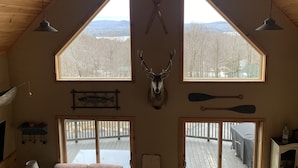 A cabin interior with a high ceiling, wooden beams, two large windows with a mountain view, a mounted deer head, and fishing equipment.
