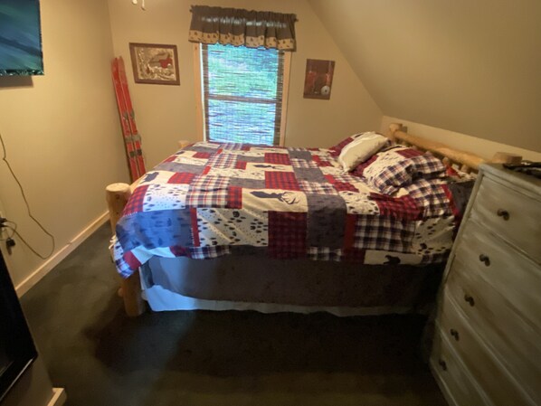 Ein Schlafzimmer mit einem mit einer Decke bezogenen Bett, einem Schminktisch, einem Fenster mit Jalousien und einem an der Wand hängenden Bild.