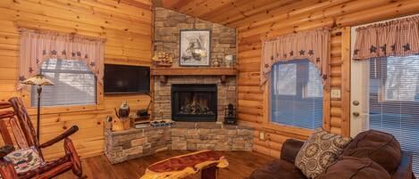 A cozy log cabin interior with a stone fireplace, wooden furniture, and a ceiling fan.