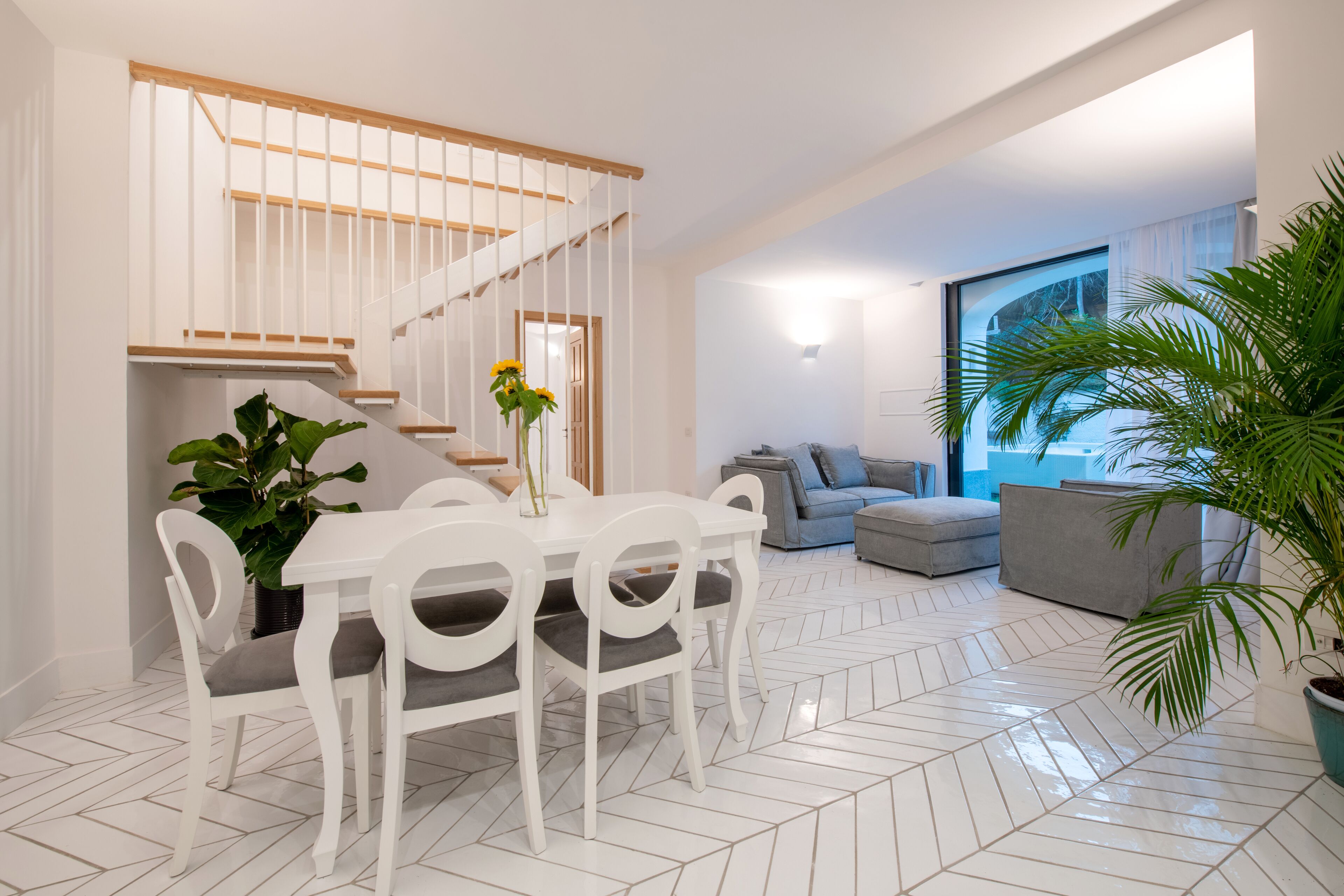 A modern living room with a white staircase, a dining area with white chairs, and a large window with a view of trees.