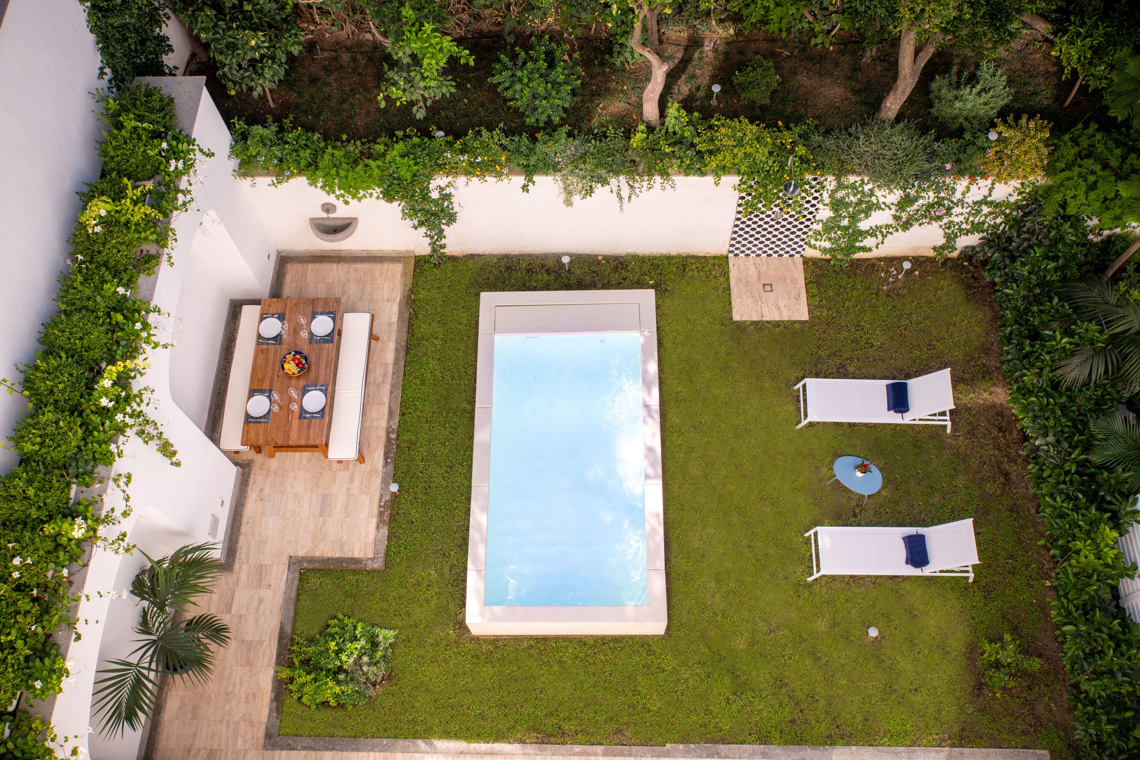 An aerial view of a backyard with a swimming pool, two lounge chairs, a dining table with chairs, and surrounding greenery.