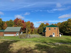 A rural property with two main buildings, one with a green roof and another with a brown exterior. There is a large rock in the foreground, and the surrounding area is grassy with trees in the background.