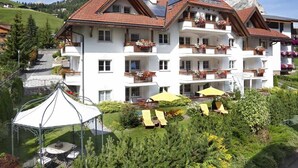 Een meerlaags hotel met balkons, een tuin met ligbedden en een berg op de achtergrond.