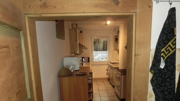 Um corredor estreito leva a uma cozinha com armários de madeira, um micro-ondas e uma janela com cortinas.