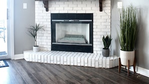 Una sala de estar con chimenea de ladrillo blanco, un televisor de pantalla plana montado encima y piso de madera oscura.