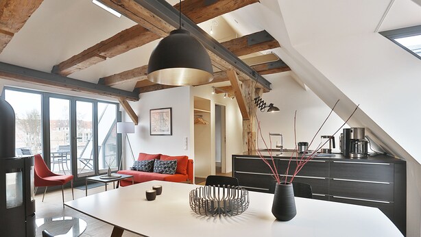 A modern loft with exposed wooden beams, a dining area with a white table and black chairs, a red sofa, and a kitchen with dark cabinetry.