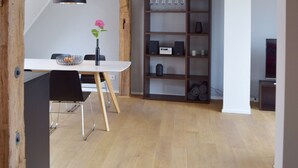 A dining area with a white table, black chairs, and a black pendant light. There is a wooden bookshelf with various items and a white pillar.