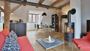A spacious living area with a sofa, a coffee table, and a fireplace. There is a kitchen in the background with a dining table and chairs.