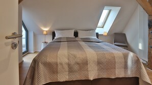 A bedroom with a sloped ceiling, a large bed, two bedside lamps, a chair, and a window.