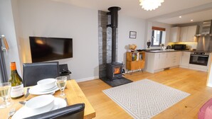 Una sala moderna con chimenea, comedor con mesa para dos y cocina con armarios blancos y electrodomésticos de acero inoxidable.