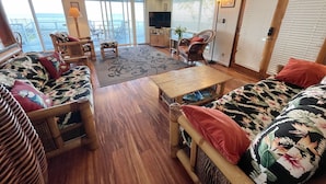 Een ruime woonkamer met een groot raam met uitzicht op de oceaan, een plafondventilator en een gezellige zithoek met gekleurde kussens.
