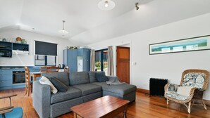 Een moderne woonkamer met een grijze modulaire bank, een houten salontafel en een rieten fauteuil. Een keuken met blauwe kastjes en een eethoek met blauwe stoelen zijn zichtbaar.