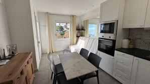 Une cuisine compacte avec des meubles blancs, une table à manger avec des chaises et une vue sur l’extérieur par une fenêtre.