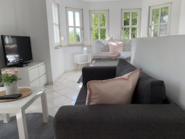 Uma sala de estar moderna com um sofá cinzento, uma TV sobre um armário branco, uma pequena mesa com um vaso de flores e uma janela com vista para o verde.
