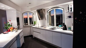A modern kitchen with white cabinetry, a built-in oven, a stainless steel range hood, a large window with a sunset view, and a refrigerator with a water dispenser. - Spacious luxury apartment with 180° ocean view,  pool, seafront beach (Parede)