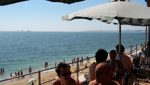 A beachside restaurant with outdoor seating overlooking the sea, a sandy beach, and a distant city skyline. - Spacious luxury apartment with 180° ocean view,  pool, seafront beach (Parede)