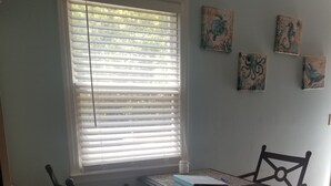 Un coin repas comprenant une table et deux chaises, une fenêtre avec des stores et des œuvres d’art murales.